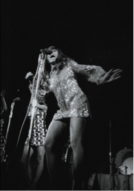 Tina Turner on stage, singing into a microphone in its stand, one arm out to her side, the other lifted but partially obscured, and wearing a long-sleeved, short sequin shift
