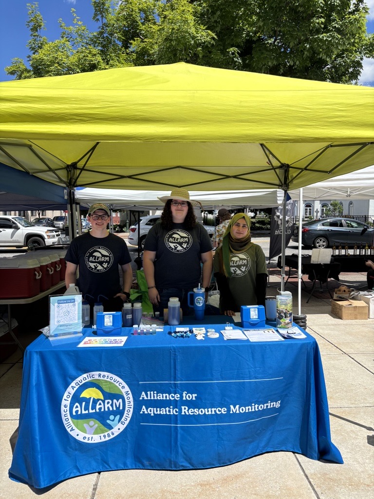 photo of Farmers on the Square tabling with Hakeema, Alyssa, and Ryan smiling behind the table. Taken from in front of the ALLARM booth.