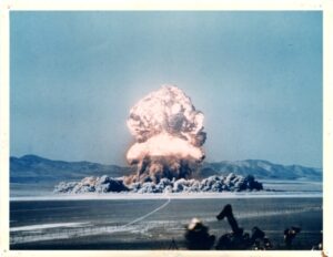 The glowing mushroom cloud of an atomic bomb at the Nevada Test Site explodes against a teal blue sky.