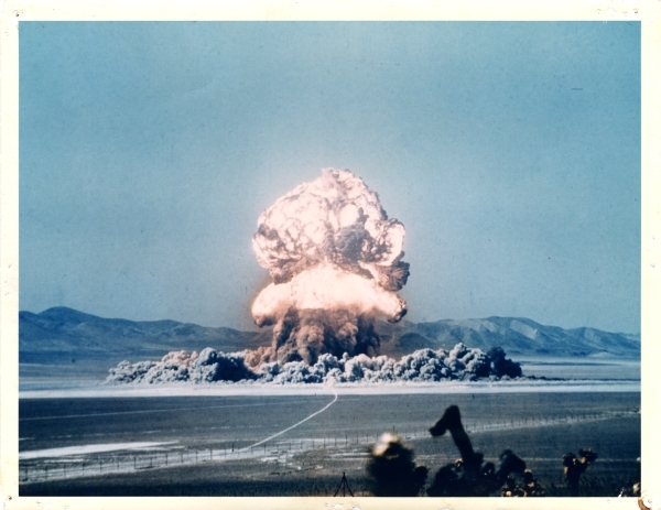 The glowing mushroom cloud of an atomic bomb at the Nevada Test Site explodes against a teal blue sky.