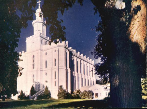 Vintage postcard photo of a large, white stone building with tall arched windows and a central tower topped by a golden statue. The structure is the St. George Utah Temple of the Church of Jesus Christ of Latter-day Saints, set against a deep blue sky and partially framed by tall trees and dark green lawn.