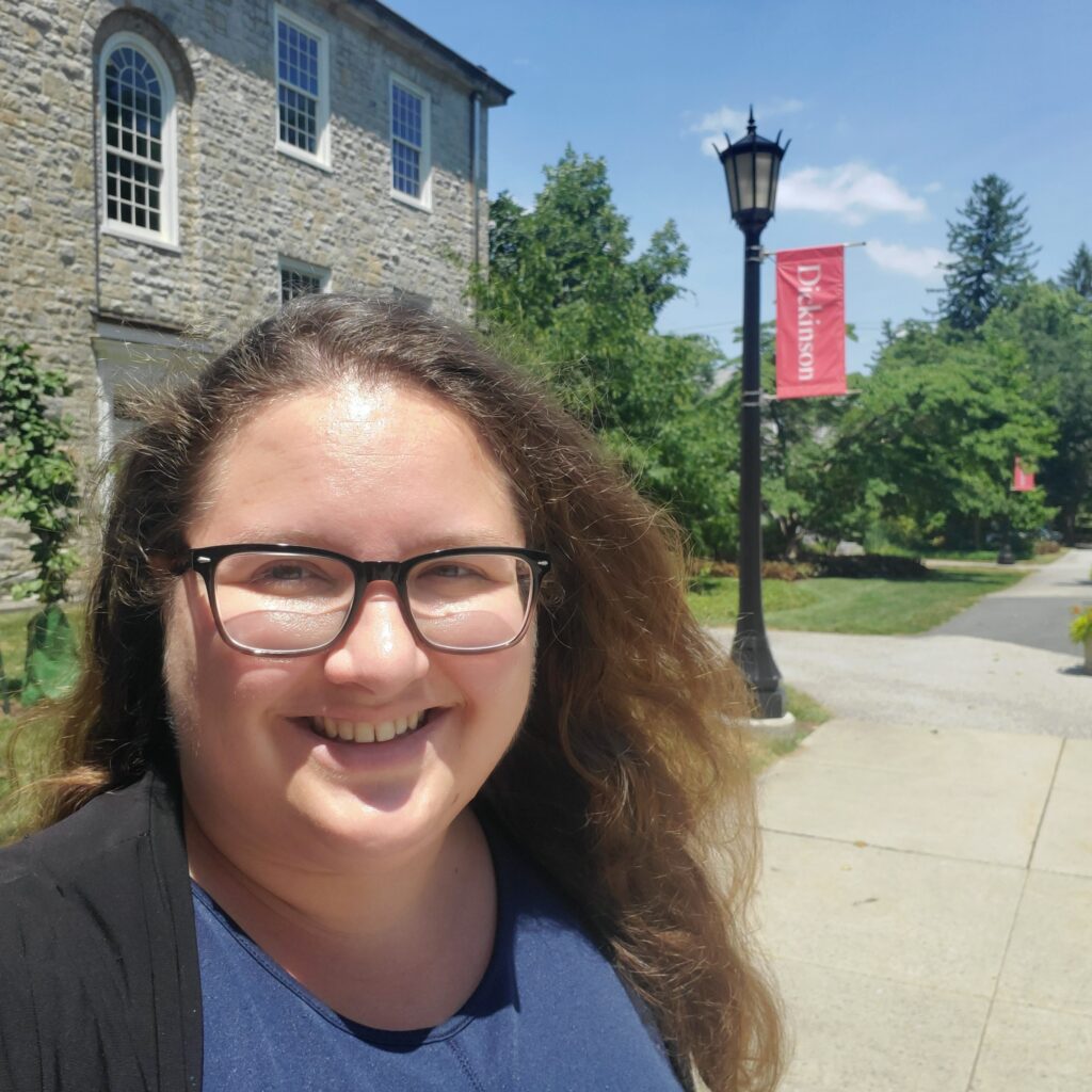 Photo of Prof. Wilkins, a white woman with brown hair (worn down) wearing black thick-rimmed glasses. Behind her is a Dickinson College campus building and a black lamp post with a red Dickinson banner.